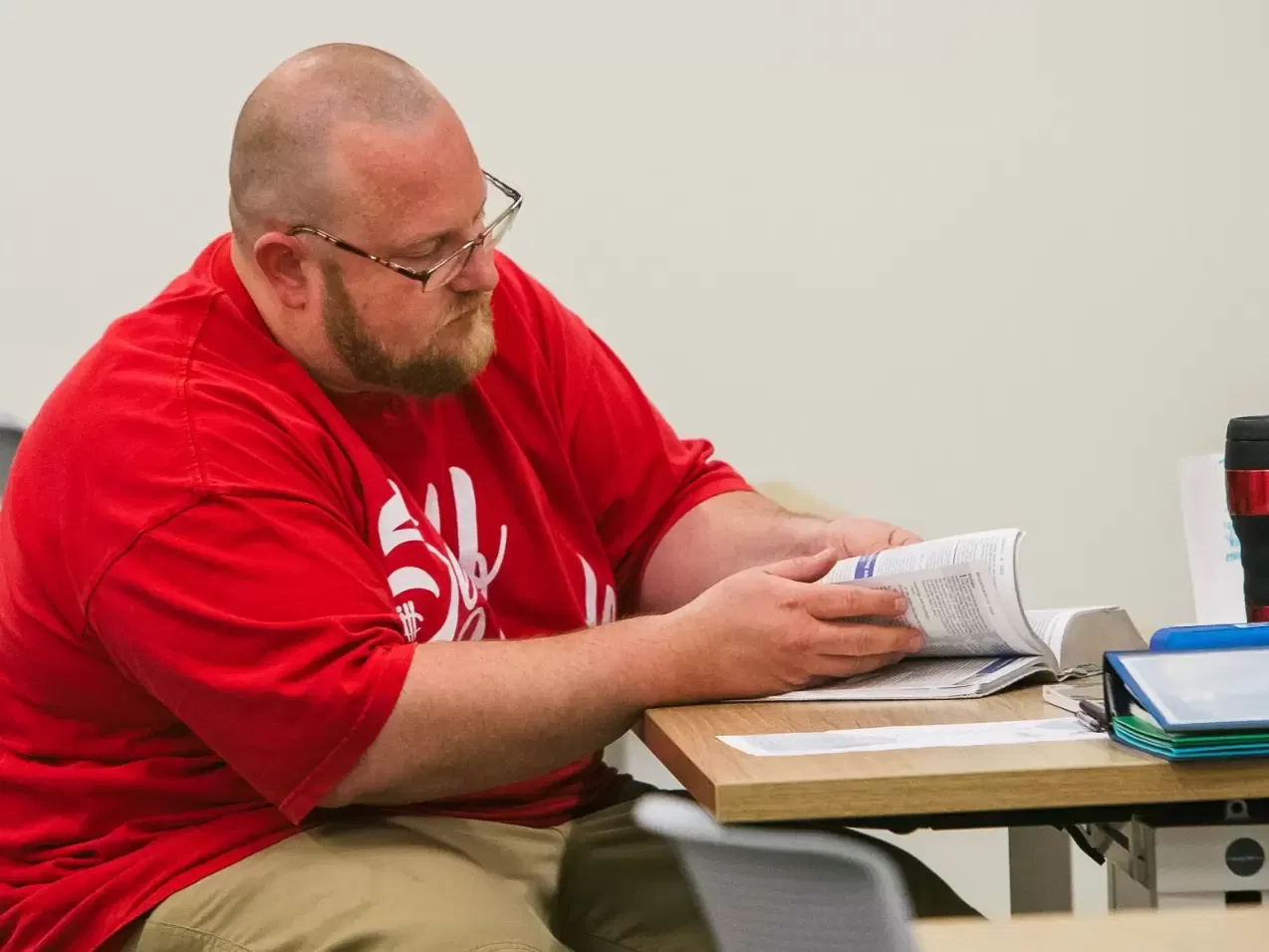 Man in a red shirt seated at a desk and reading a book